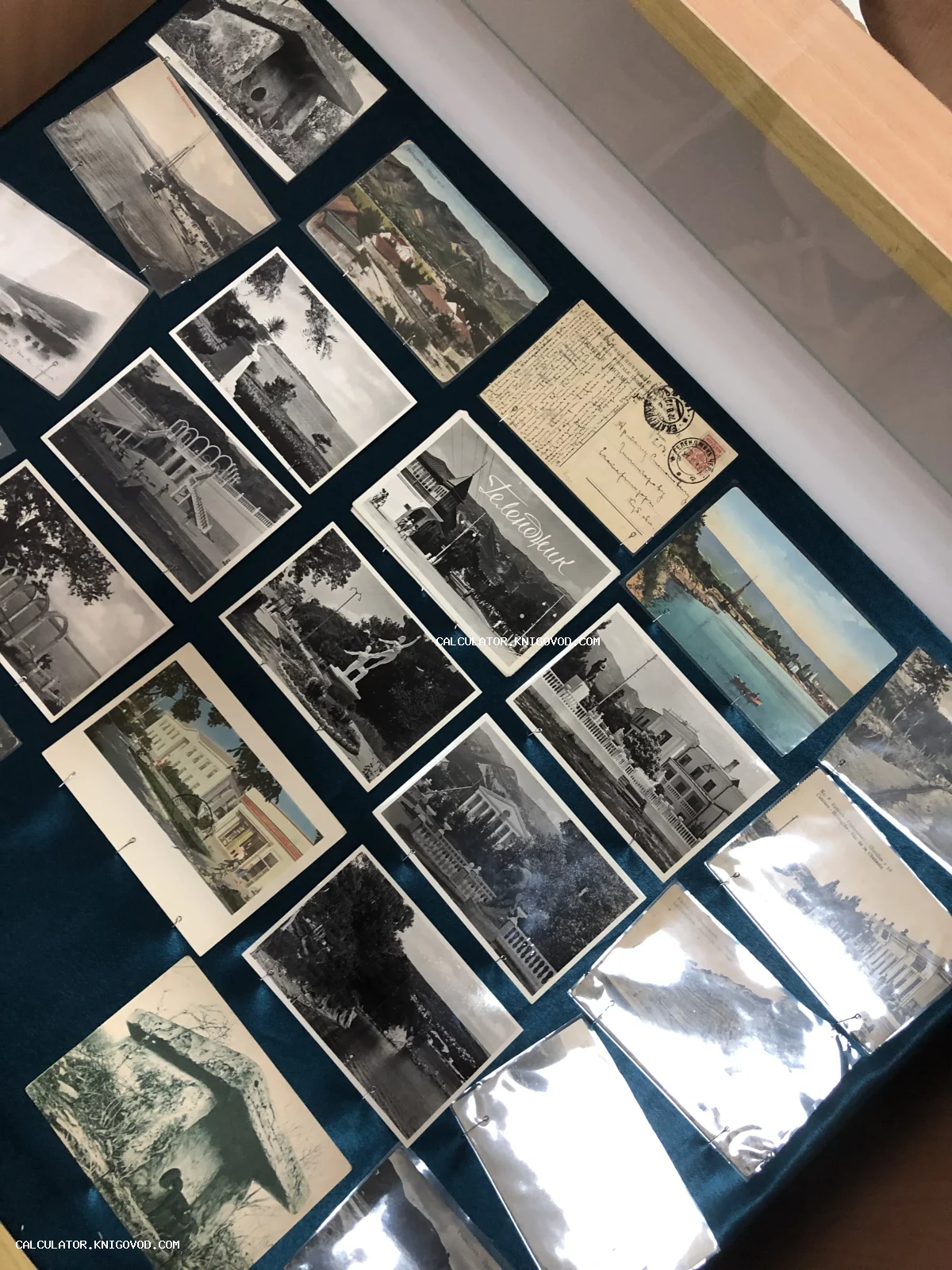 Множество старинных черно-белых и цветных открыток с изображениями архитектуры, памятников и природы южных курортов, разложенных на синем фоне под стеклом.