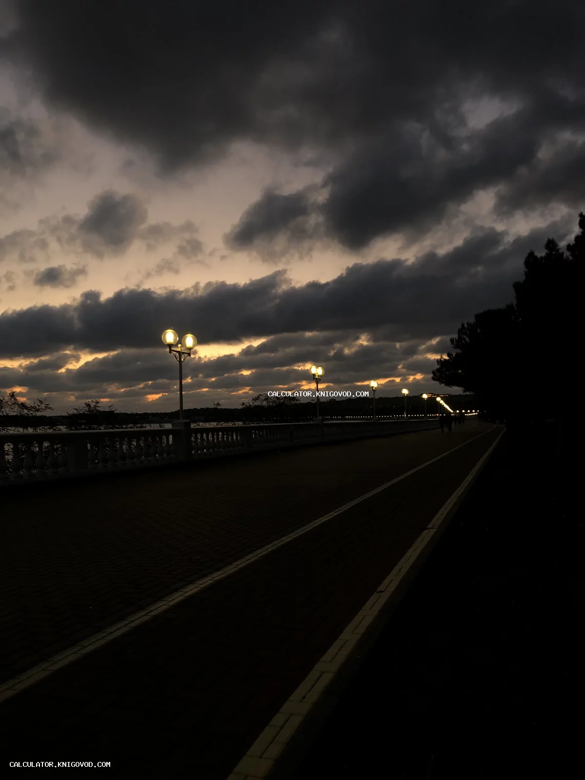 Ночная набережная с классическими круглыми фонарями вдоль балюстрады под драматичным облачным небом на закате.