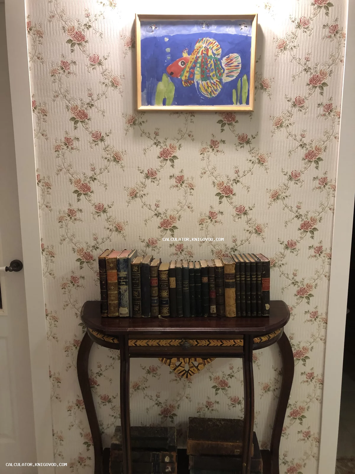 Ряд старинных книг в кожаных и тканевых переплетах на деревянном столике-консоли на фоне обоев с цветочным принтом.