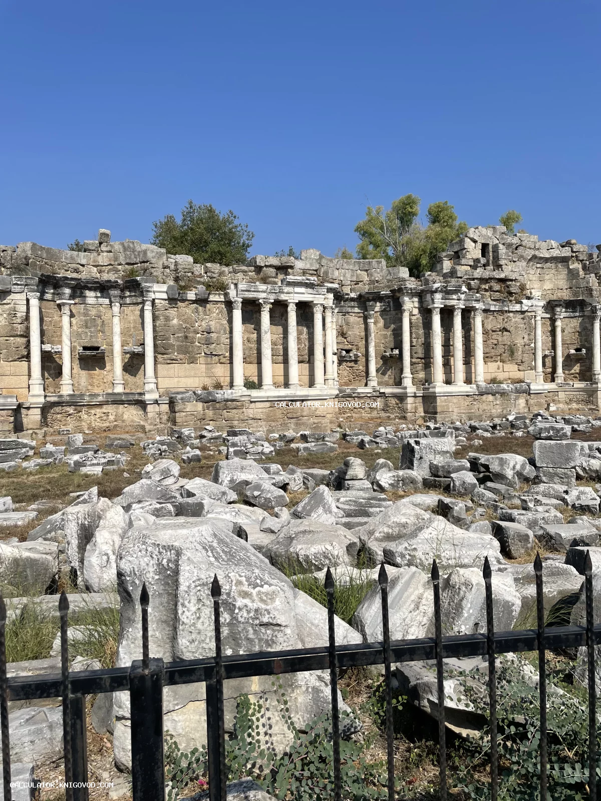 Руины античного монументального фонтана Нимфеума с белыми мраморными колоннами под ярко-синим небом.