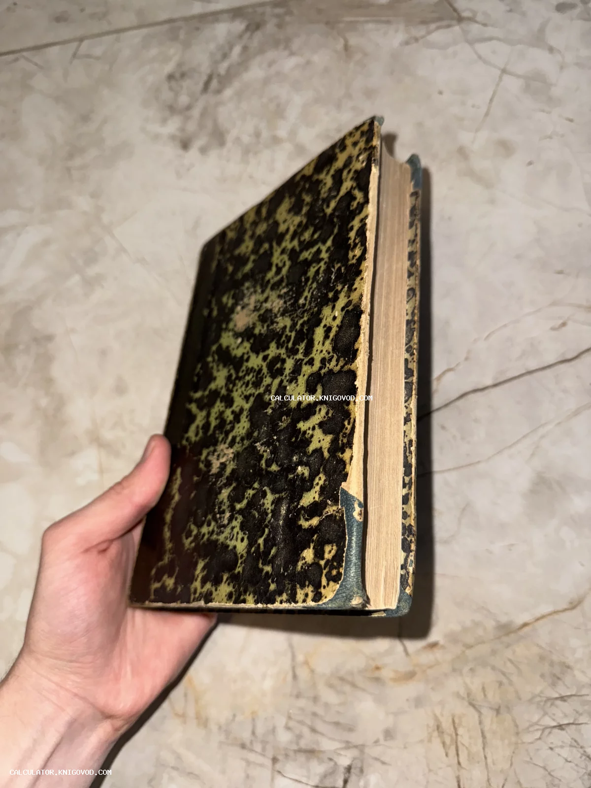 Старинная книга с характерным пятнистым мраморным узором на обложке, которую держат в руке на фоне мраморного пола.