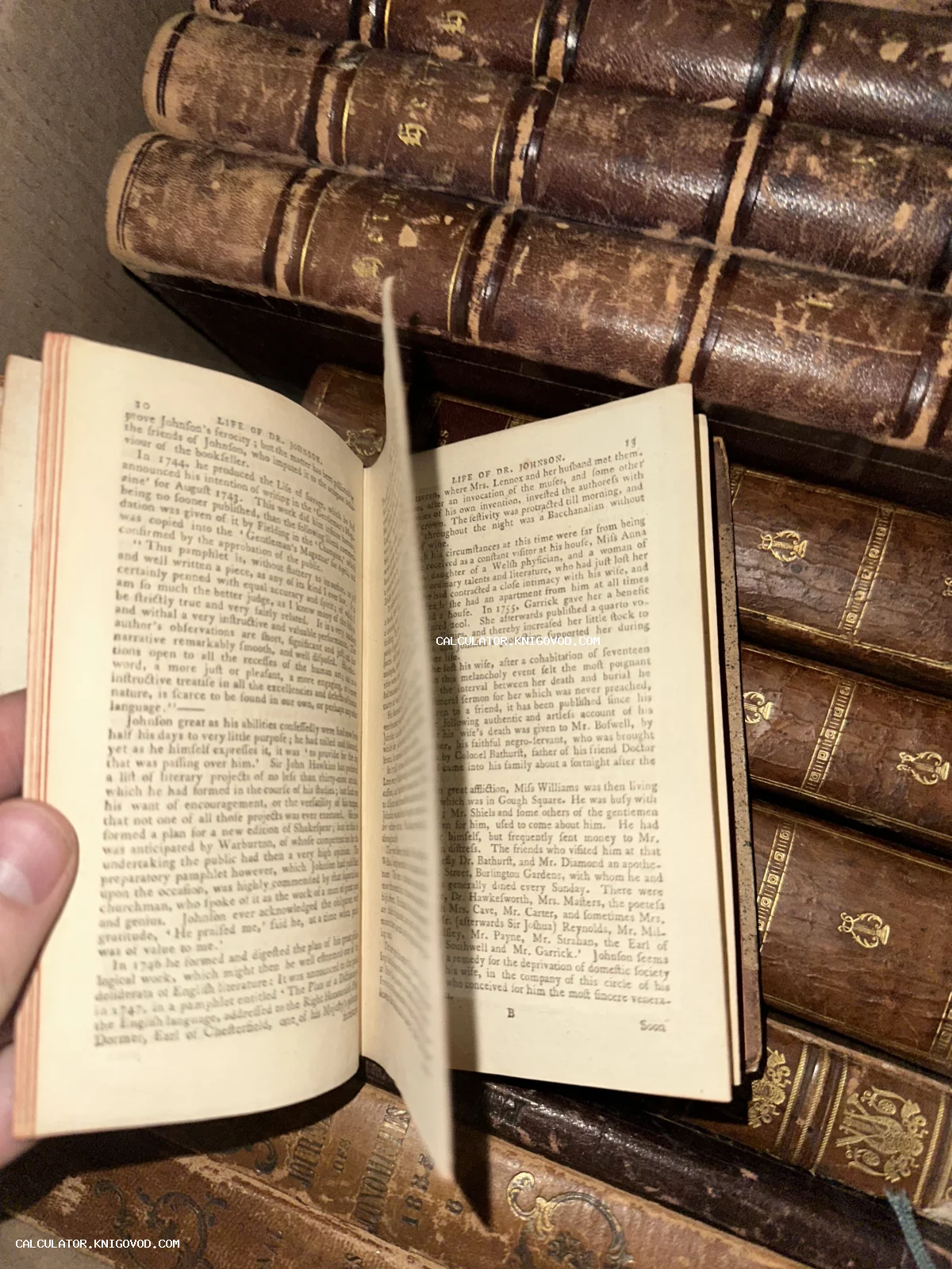 Раскрытая старинная книга с английским текстом на фоне стопки антикварных томов в кожаных переплетах с золотым тиснением.