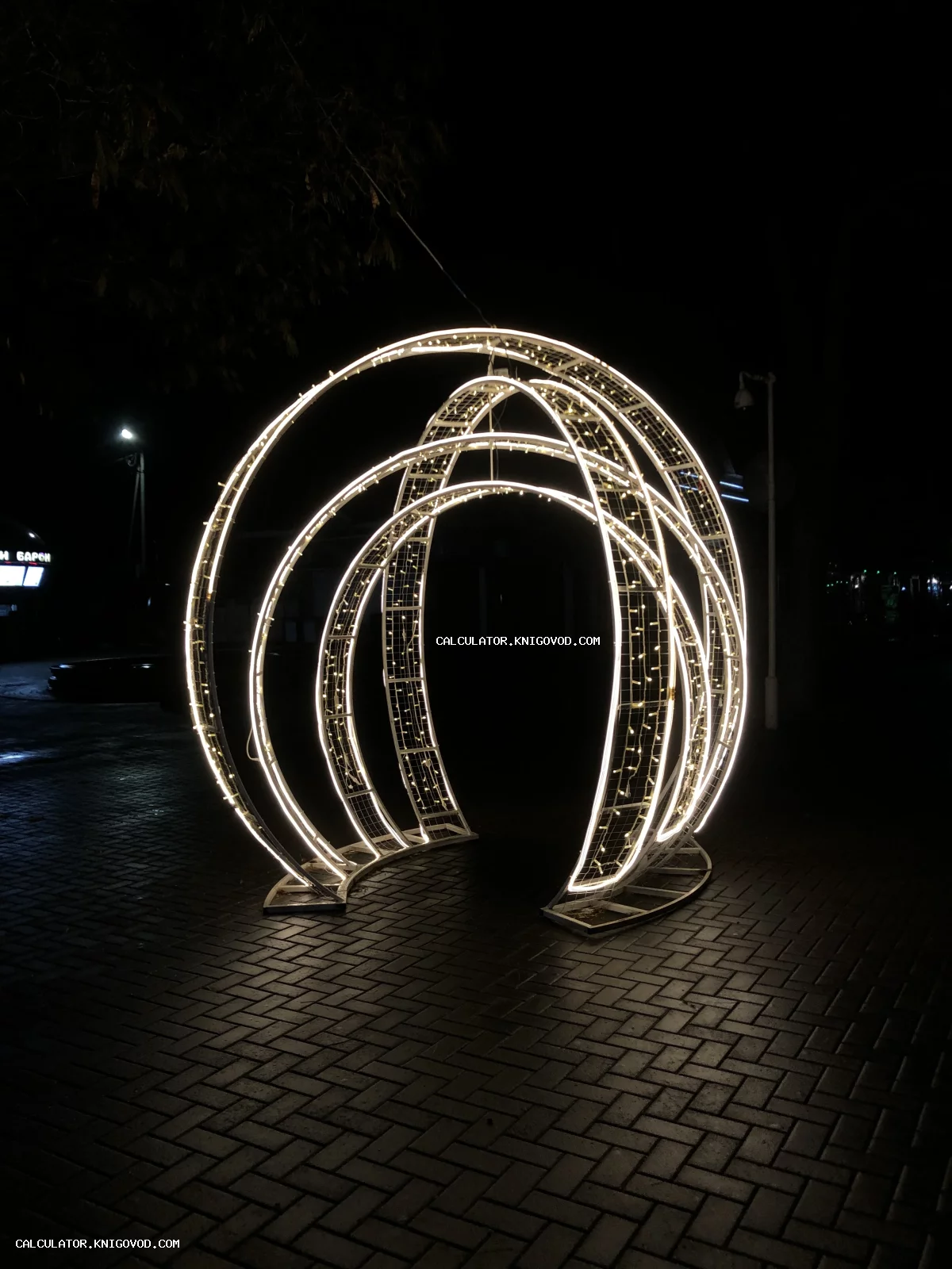 Светящаяся арт-инсталляция из нескольких белых арок на фоне темной мощеной площади ночью.