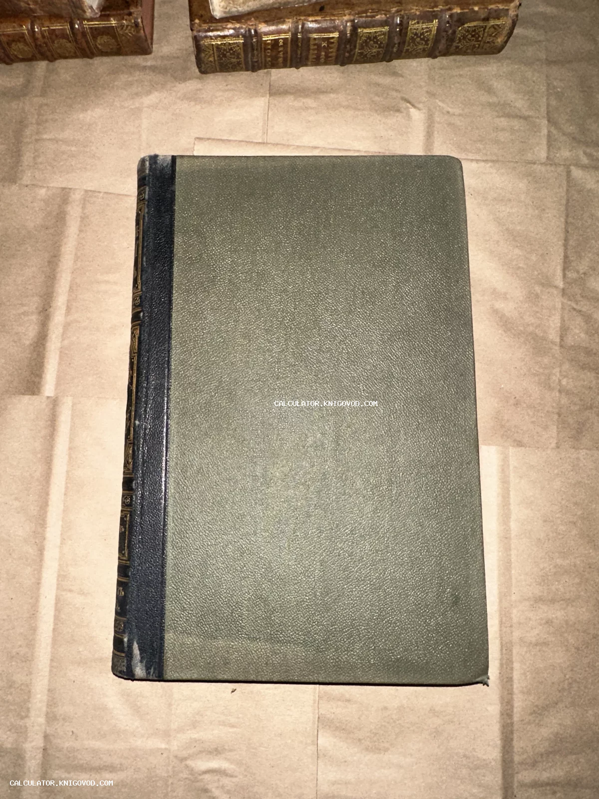 Вид сверху на антикварную книгу с зеленой фактурной обложкой и темным кожаным корешком с золотым тиснением на фоне упаковочной бумаги.