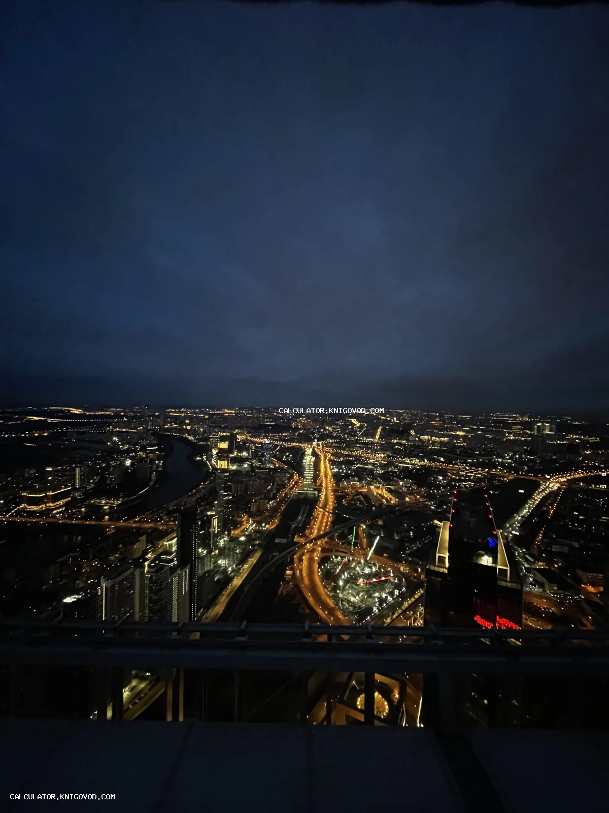 Панорамный вид на ночной город с большой высоты, светящиеся дорожные развязки, изгиб реки и огни зданий под темным небом.