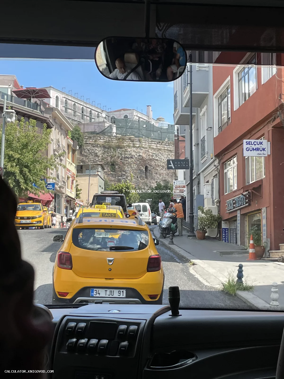 Вид из окна автомобиля на узкую городскую улицу в Стамбуле с желтыми такси и массивной древней каменной стеной на фоне.