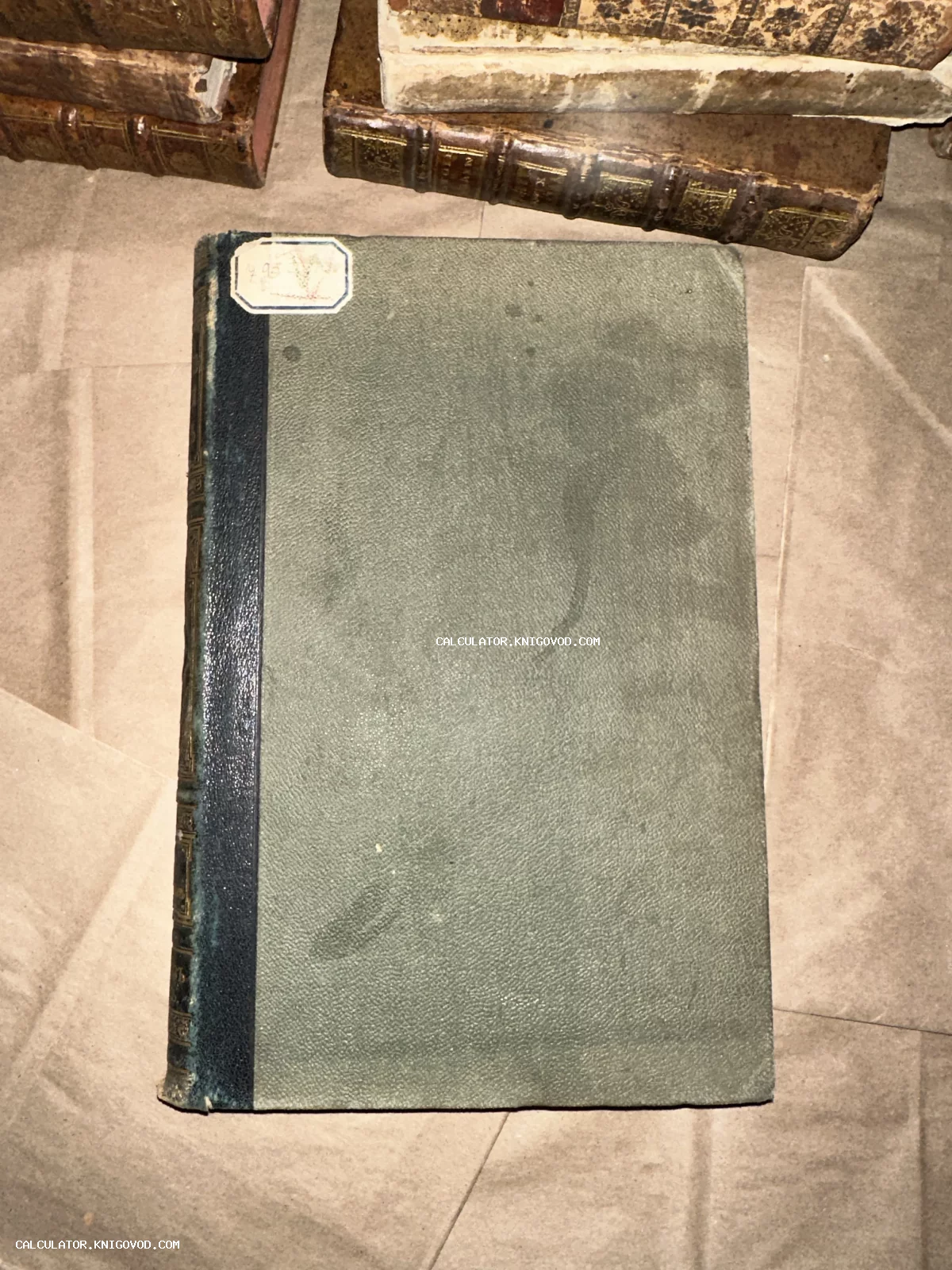 Антикварная книга с серой текстурированной обложкой и темным кожаным корешком на фоне других старых томов.