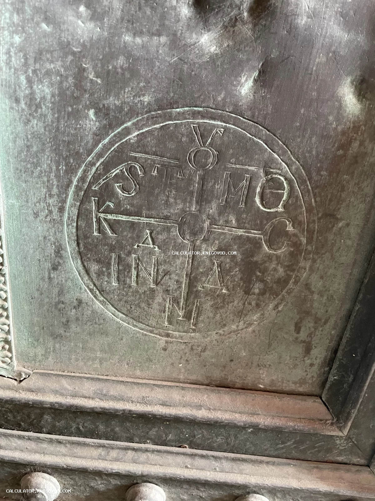 Круглая гравировка с греческими или церковнославянскими буквами на старинной металлической поверхности с патиной.