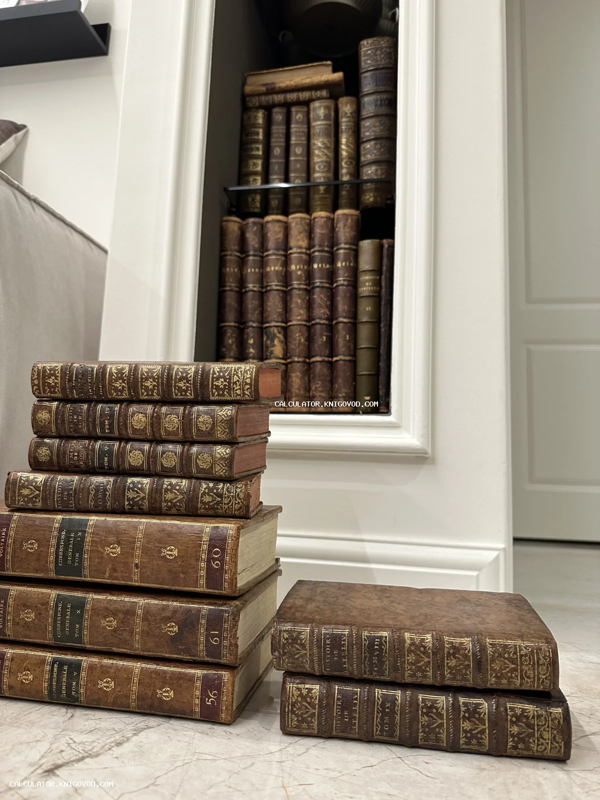 Старинные книги в кожаных переплетах с золотым тиснением, сложенные стопками на фоне белой ниши с книжными полками.