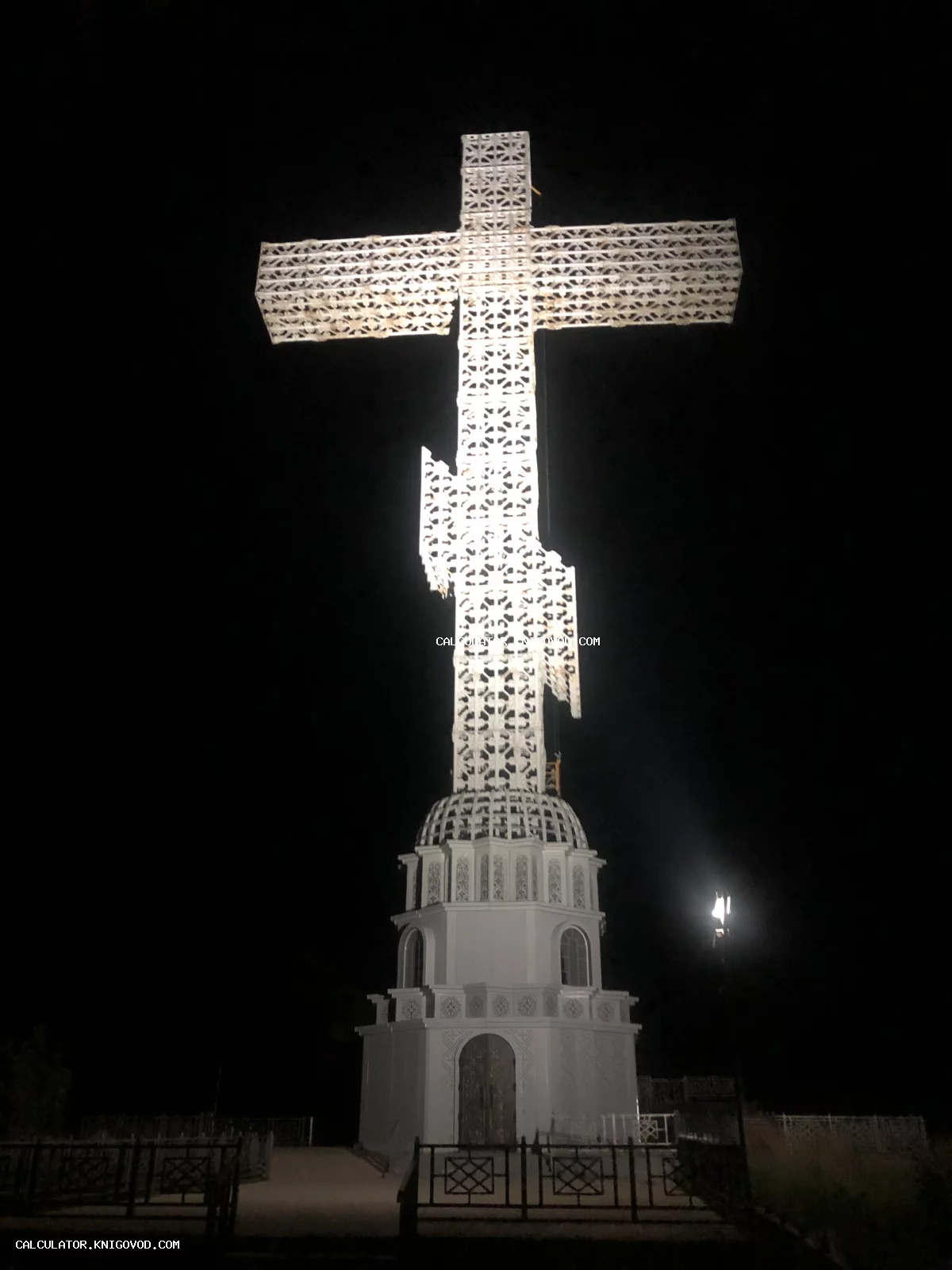 Высокий белый крест с ажурными узорами на массивном белом основании, ярко подсвеченный на фоне черного ночного неба.