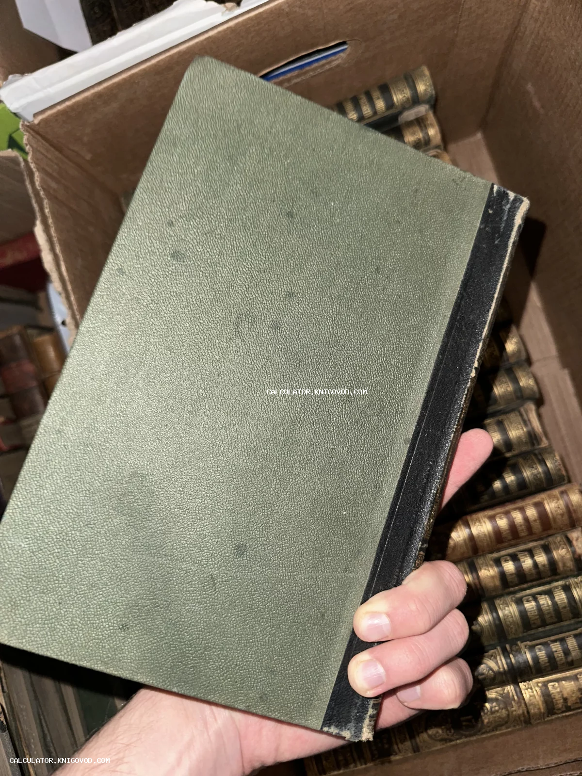 Старинная книга с зеленой фактурной обложкой и черным корешком в руке на фоне коллекции книг.