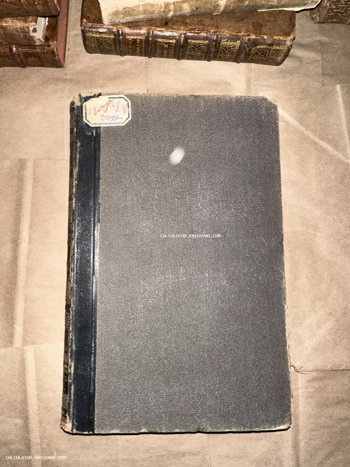 Старинная книга с темной текстурированной обложкой и бумажным ярлыком в углу на фоне других книг.