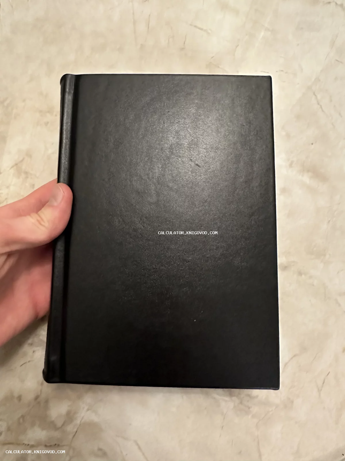 Вид сверху на черную книгу в твердом переплете с текстурой кожи, которую придерживает рука на фоне светлого мрамора.