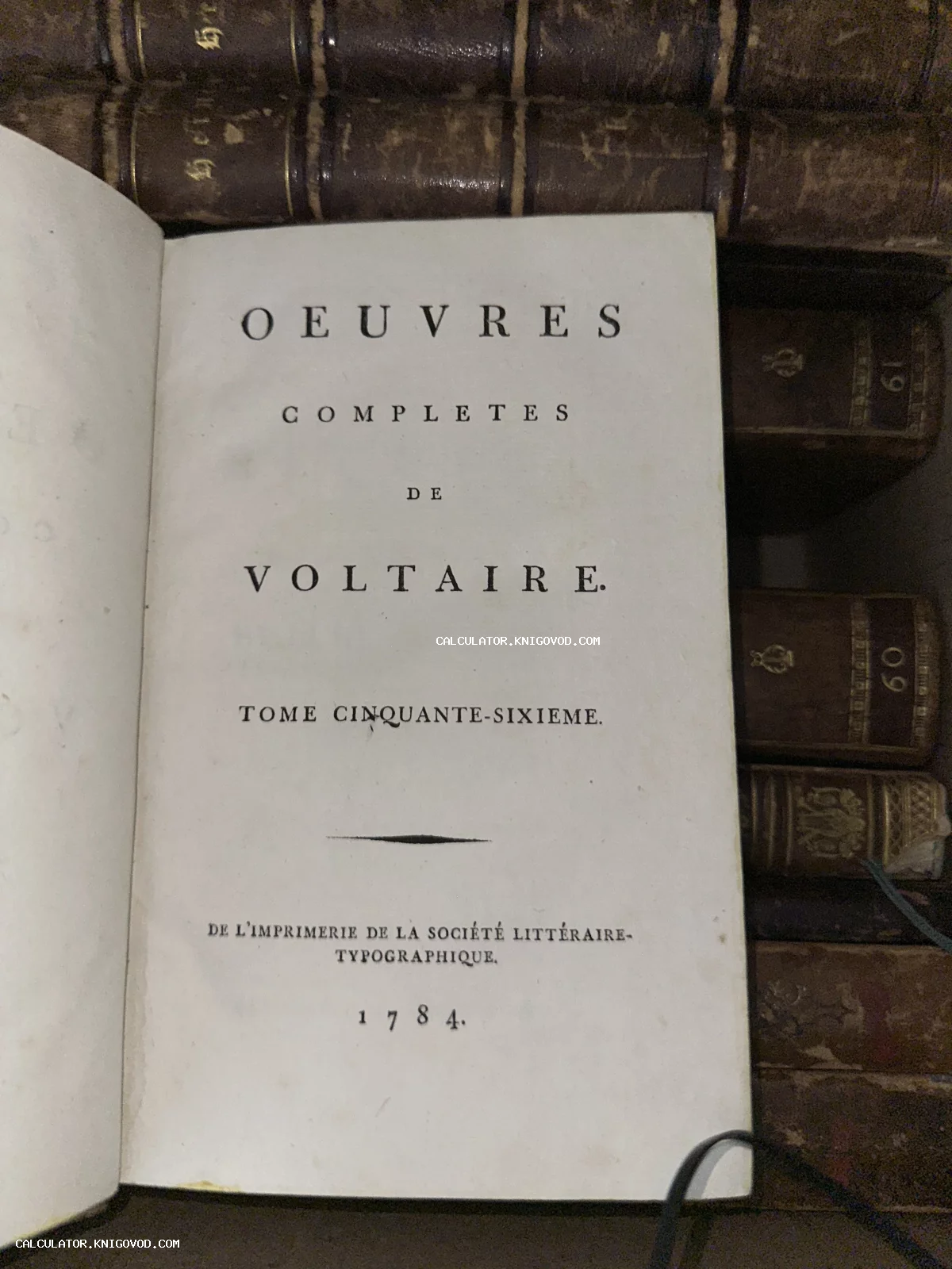 Титульный лист антикварной французской книги Вольтера Oeuvres Completes 1784 года издания на фоне старинных кожаных переплетов.