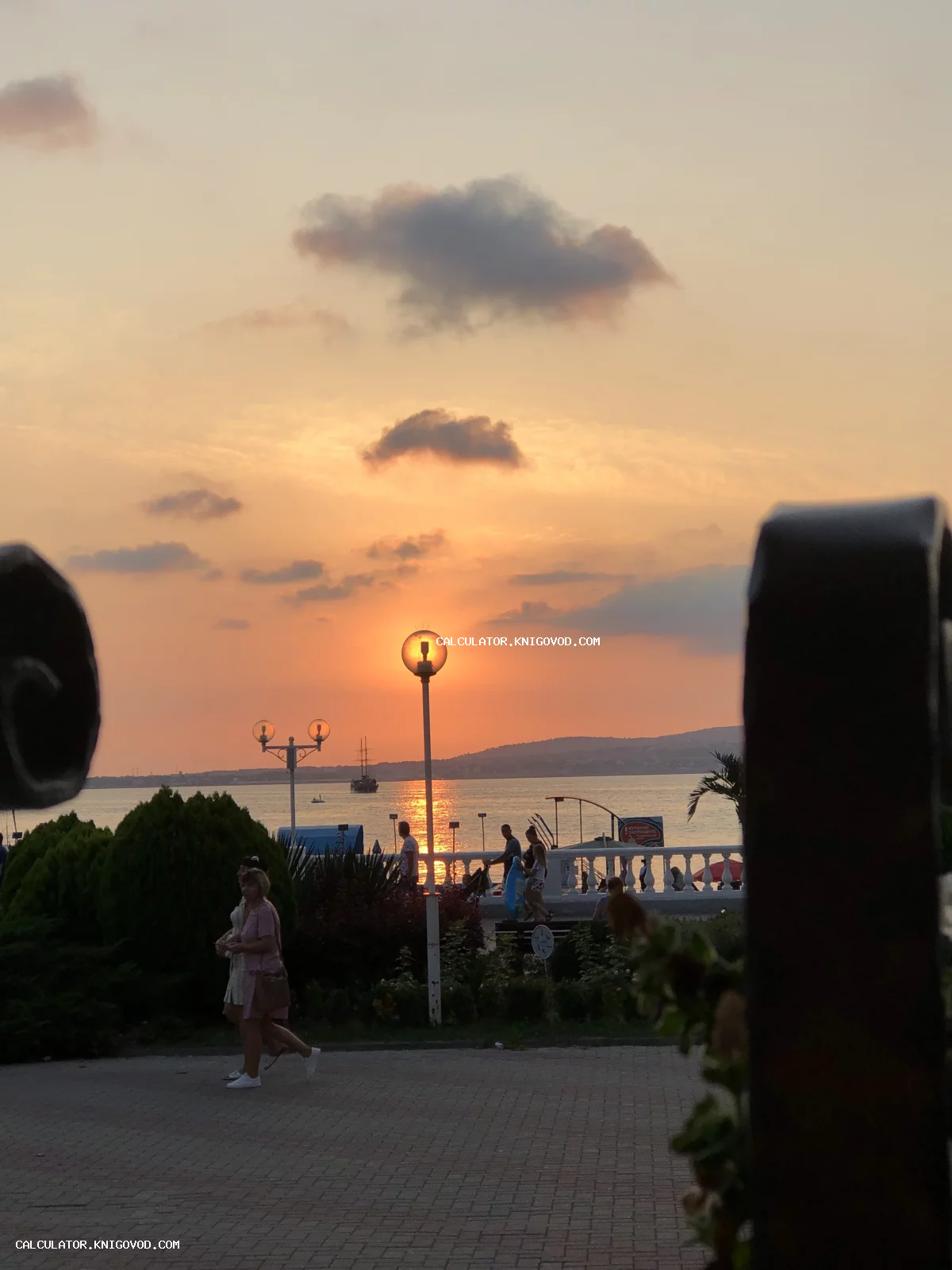 Закат над морской бухтой: набережная с белой балюстрадой, фонари и гуляющие люди в лучах заходящего солнца.