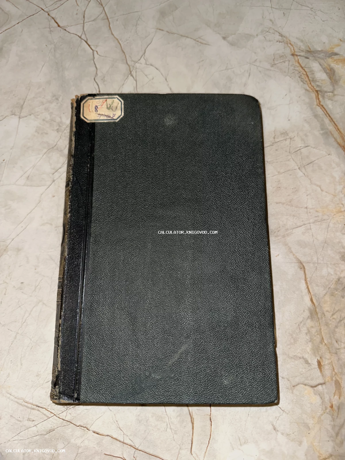 Старинная книга с черной фактурной обложкой и небольшой бумажной этикеткой в верхнем углу на фоне мраморной поверхности.
