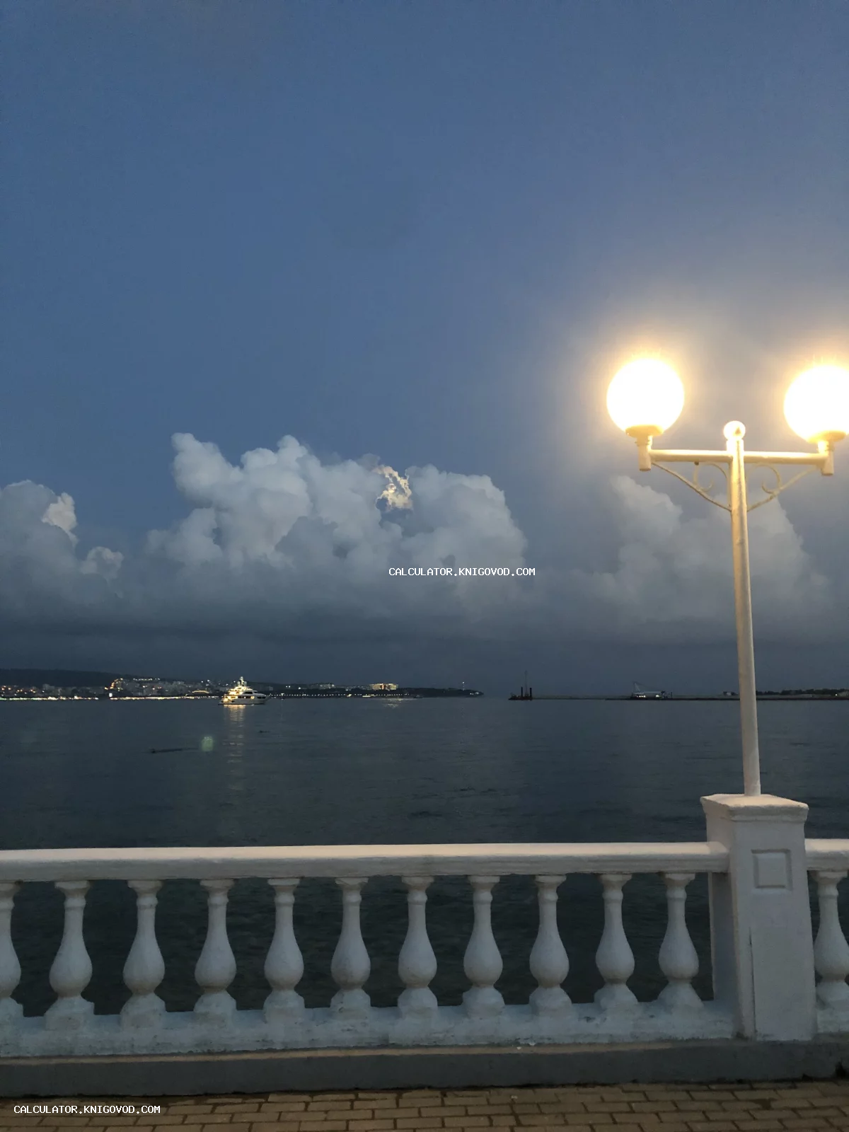 Вид на морскую бухту в сумерках с белой балюстрадой и горящим уличным фонарем на переднем плане.