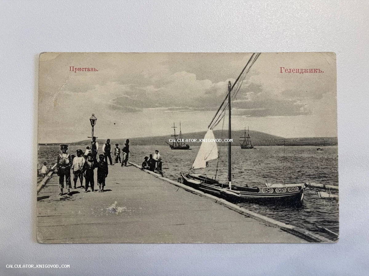 Старинная черно-белая открытка с видом на пристань в Геленджике, людьми на причале и парусными судами на фоне горного побережья.