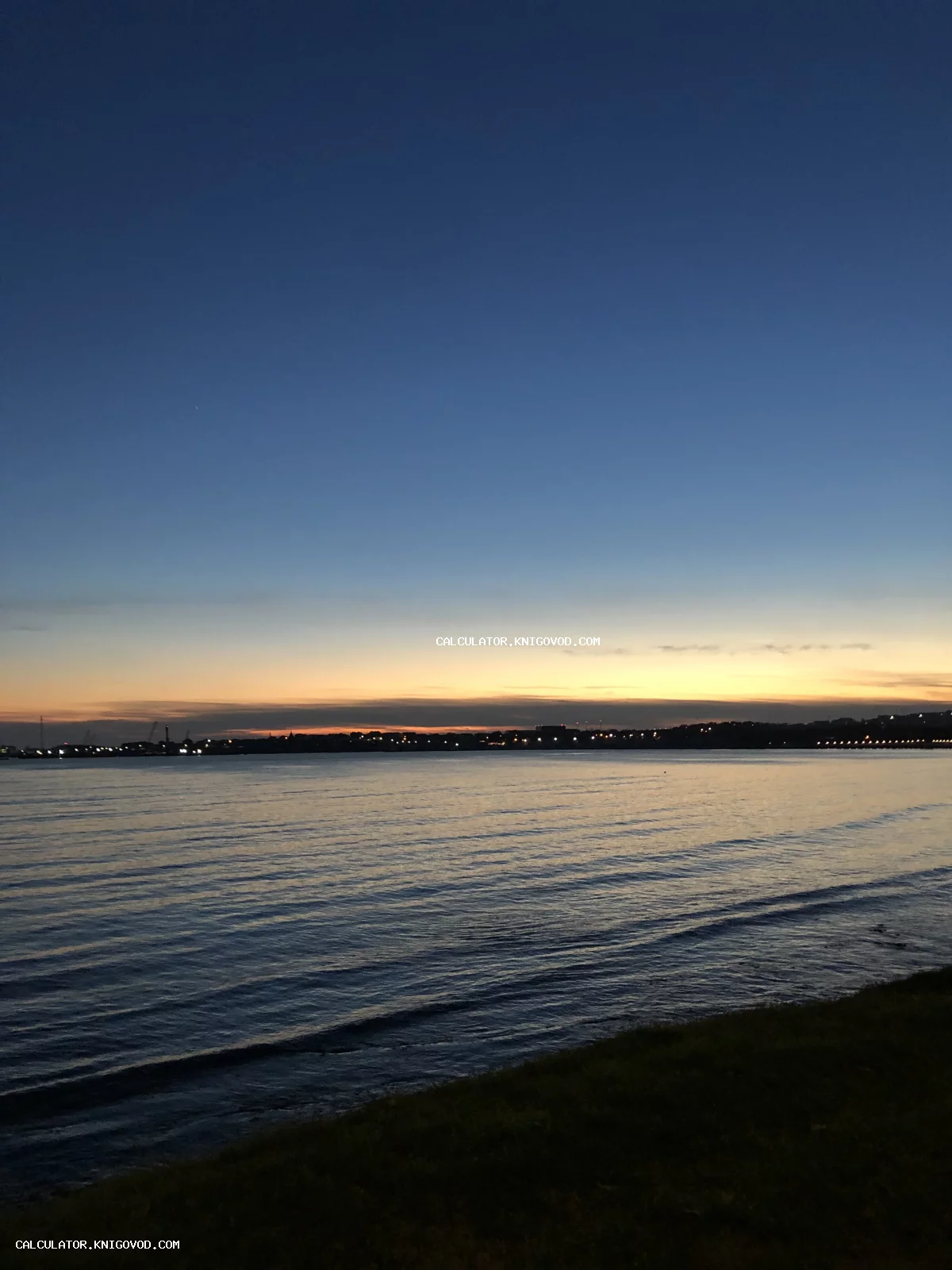 Закат над спокойным морем с огнями города на горизонте и глубоким синим небом.
