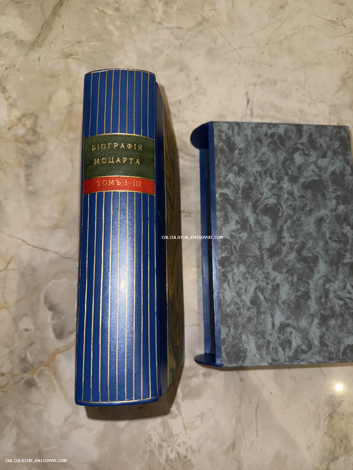 Корешок старинной книги «Биография Моцарта» в синем кожаном переплете с золотым тиснением и защитный футляр на мраморном фоне.