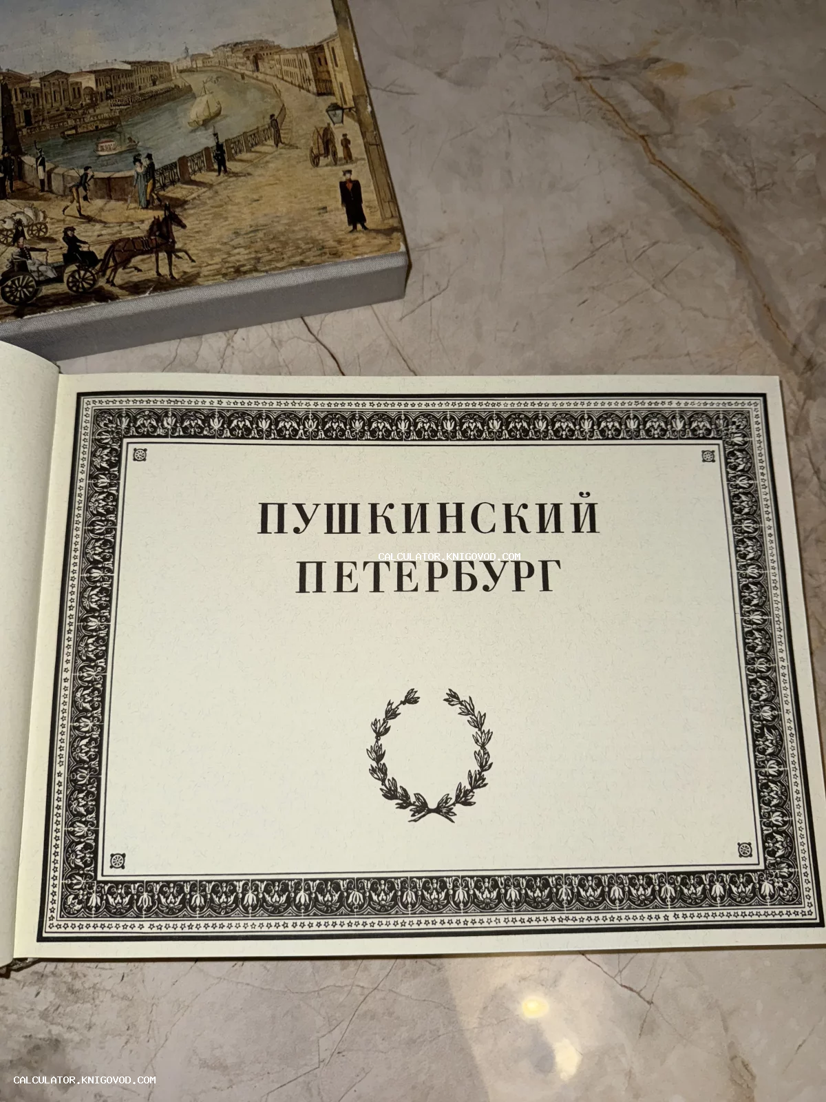 Титульный лист антикварного альбома «Пушкинский Петербург» с классическим орнаментом и лавровым венком, рядом виден иллюстрированный футляр.