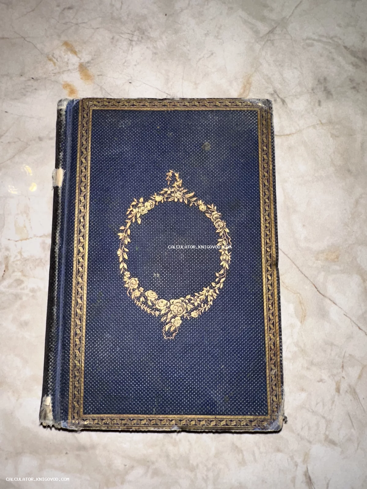 Старинная книга в темно-синем переплете с золотым тиснением в виде цветочного венка и декоративной рамки.