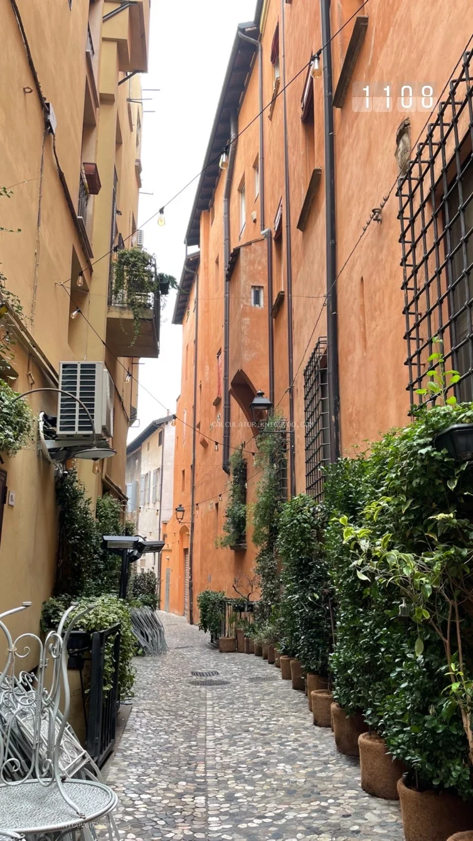 Узкая мощеная улица между высокими зданиями персикового цвета с обилием зеленых растений в горшках и кованой мебелью на переднем плане.