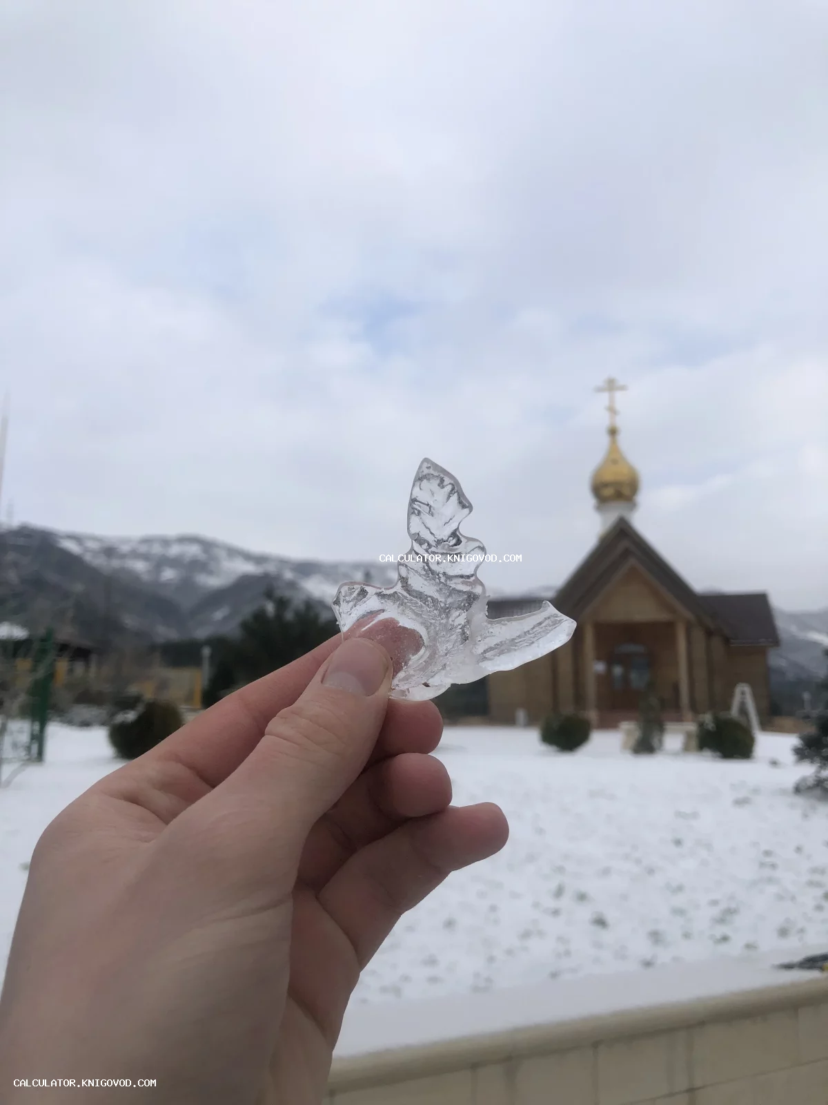 Рука держит прозрачный осколок льда в форме ангела на фоне деревянной церкви и заснеженных гор.