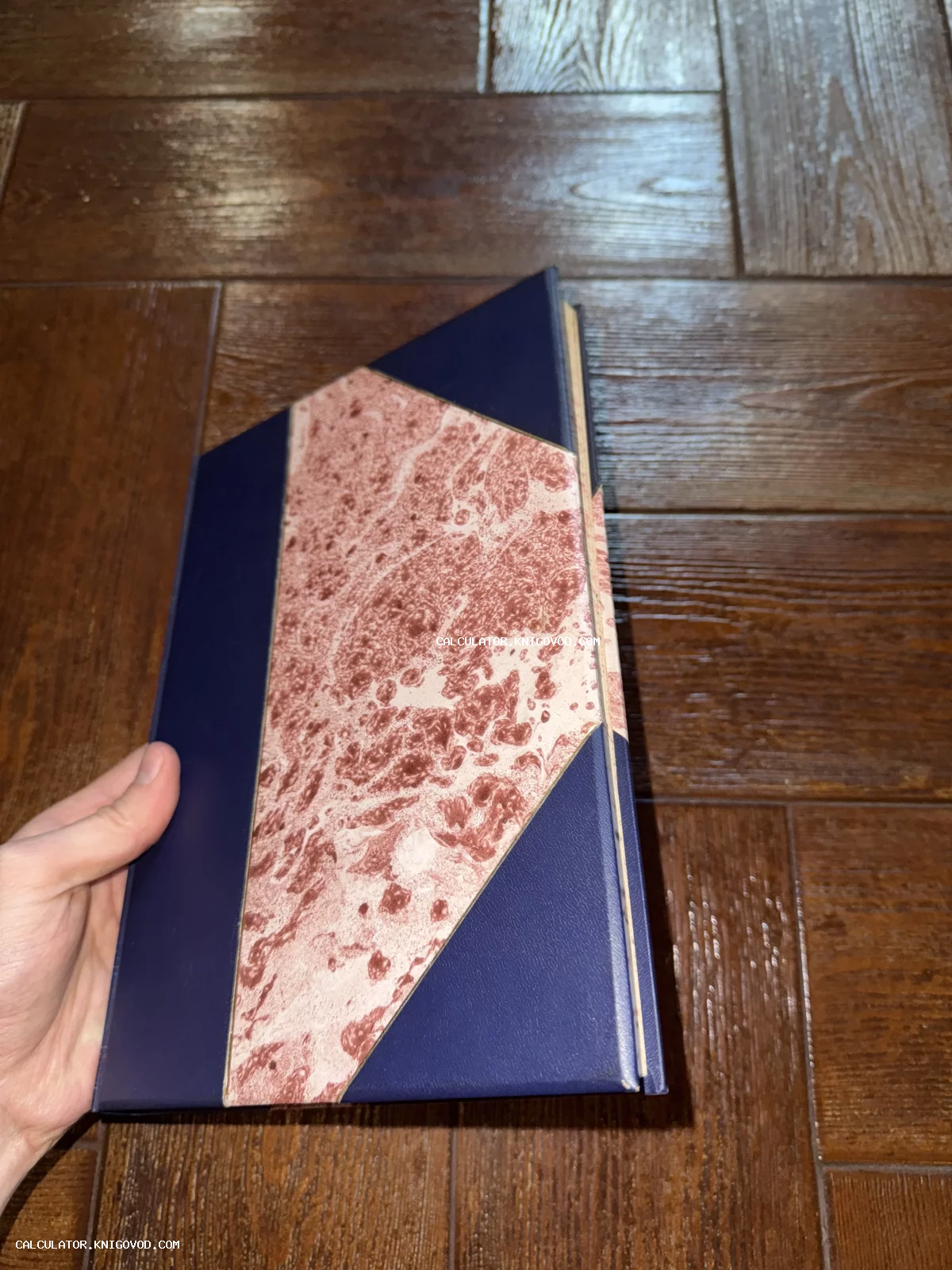 Книга в твердом темно-синем переплете с розово-коричневой мраморной бумагой на обложке на фоне деревянного пола.