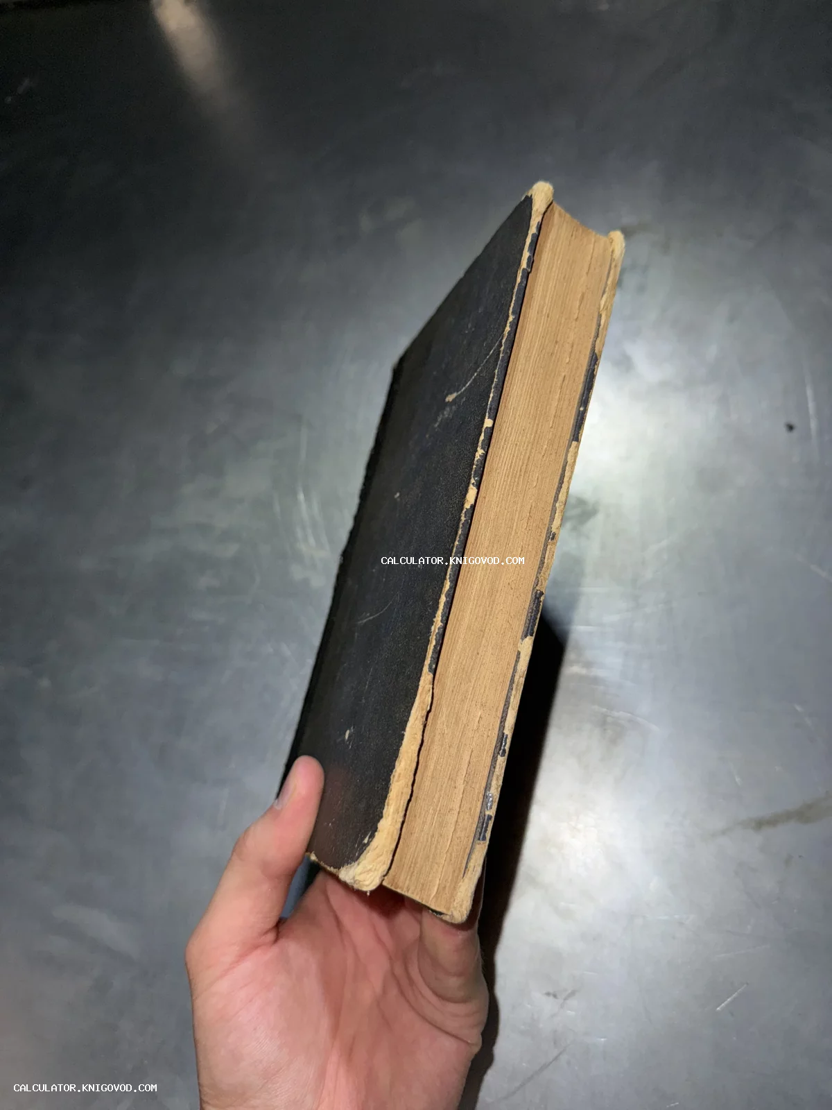 Рука держит старую книгу с пожелтевшими от времени страницами и потертой черной обложкой на темном фоне.