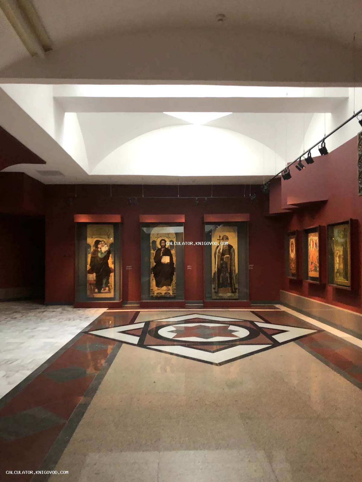 Интерьер музейного зала с красными стенами, старинными иконами в стеклянных витринах и сложной мраморной мозаикой на полу.