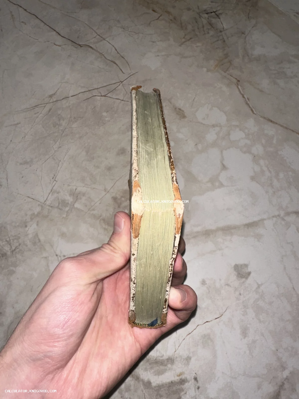 Мужская рука держит старинную малоформатную книгу, демонстрируя плотный книжный блок и потертый переплет на фоне мраморной поверхности.