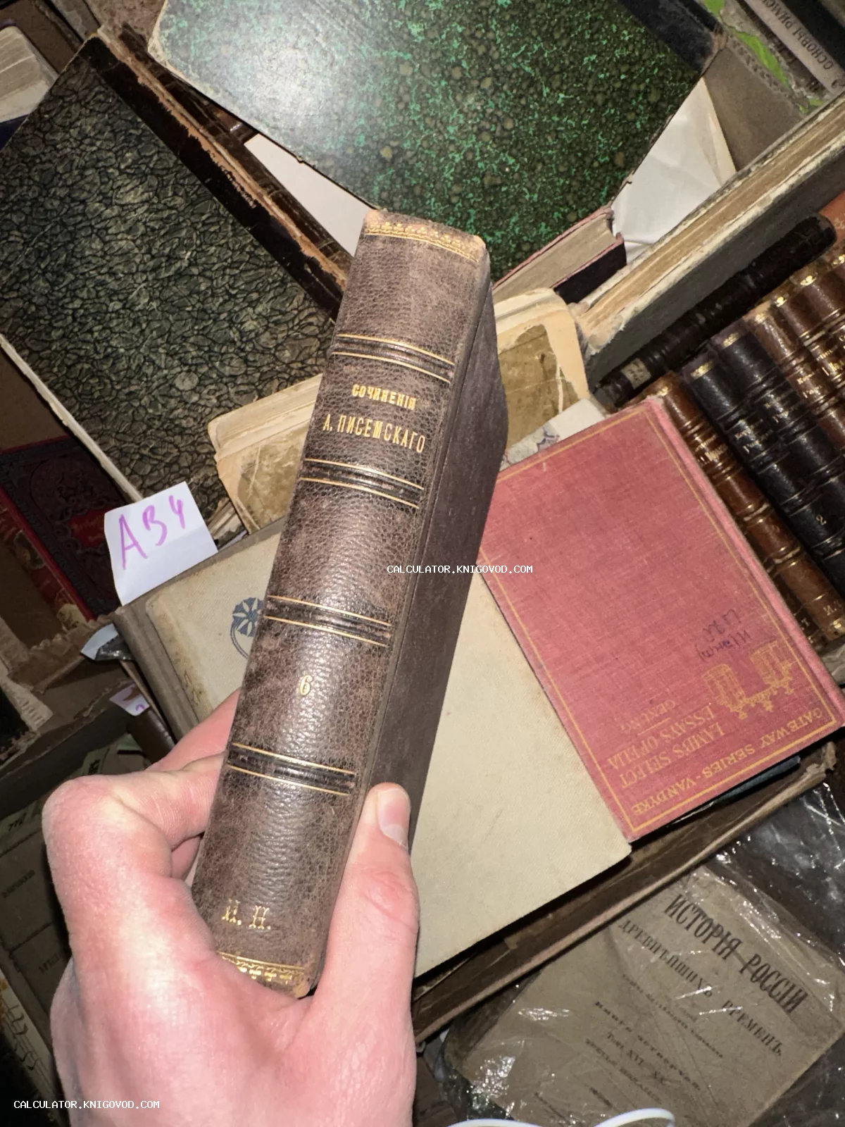 Старинная книга с кожаным корешком и золотым тиснением 'Сочинения А. Писемскаго' в руках человека на фоне других антикварных томов.