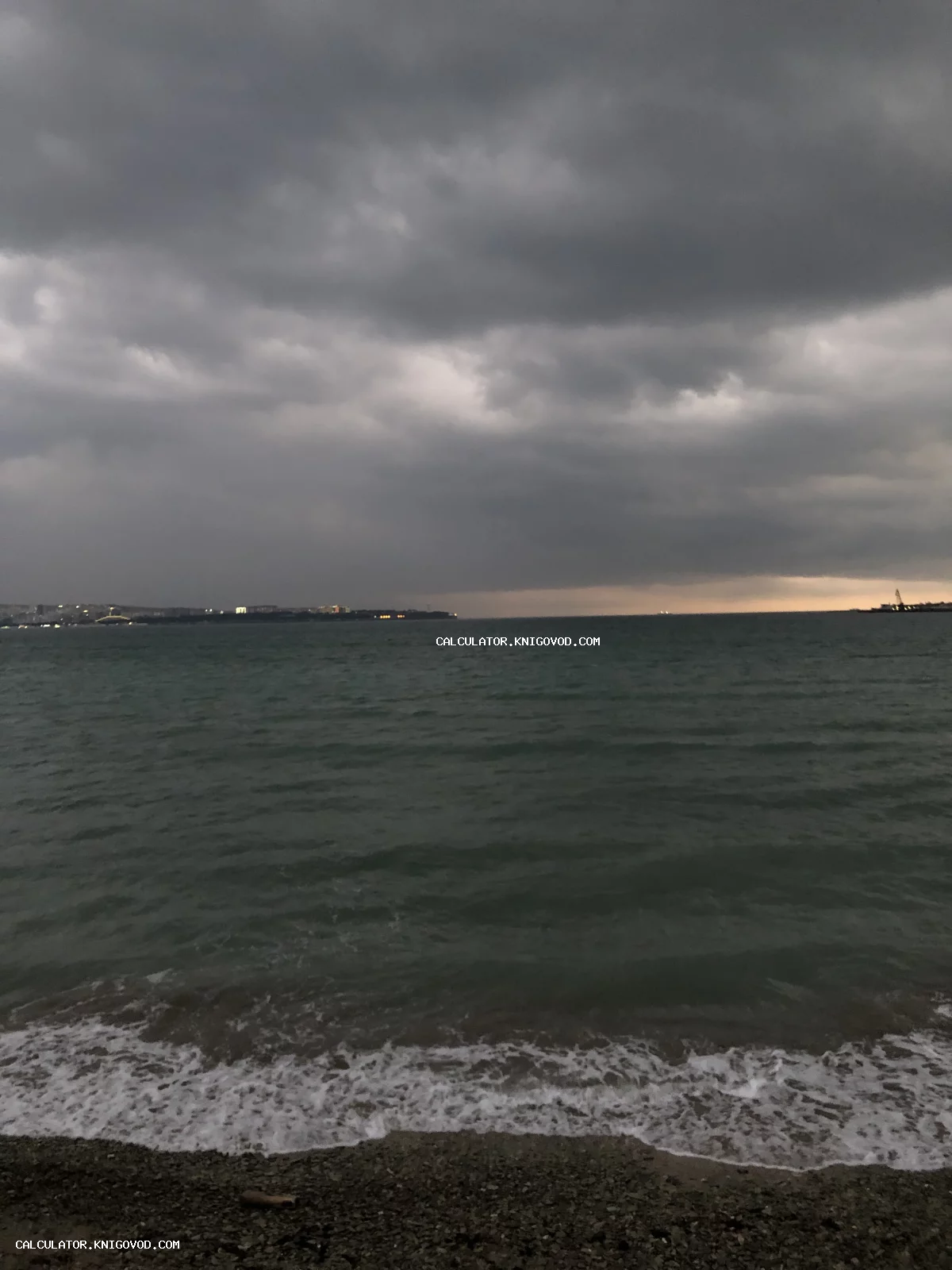 Вид на темное неспокойное море под тяжелыми грозовыми тучами, белая пена прибоя на переднем плане и огни города на горизонте.