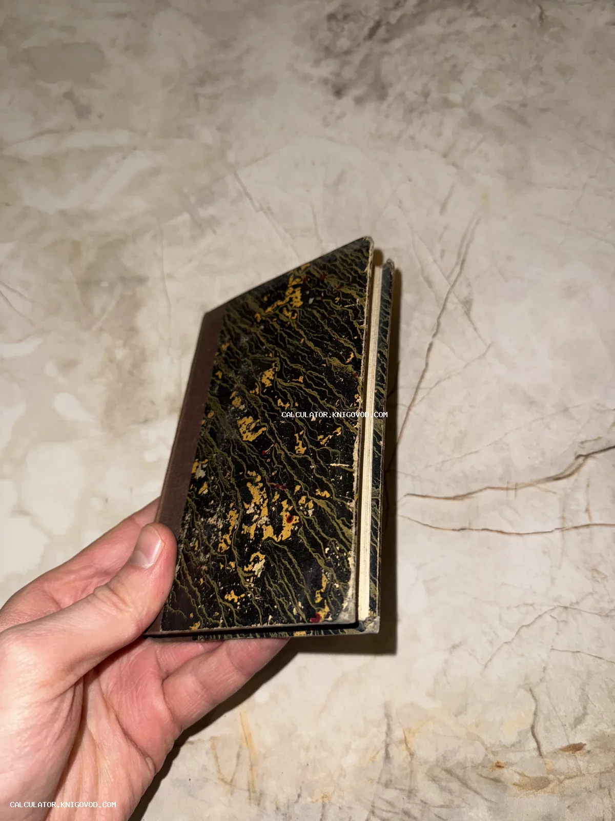 Маленькая старинная книга с обложкой из мраморной бумаги в руке человека на фоне светлого камня.