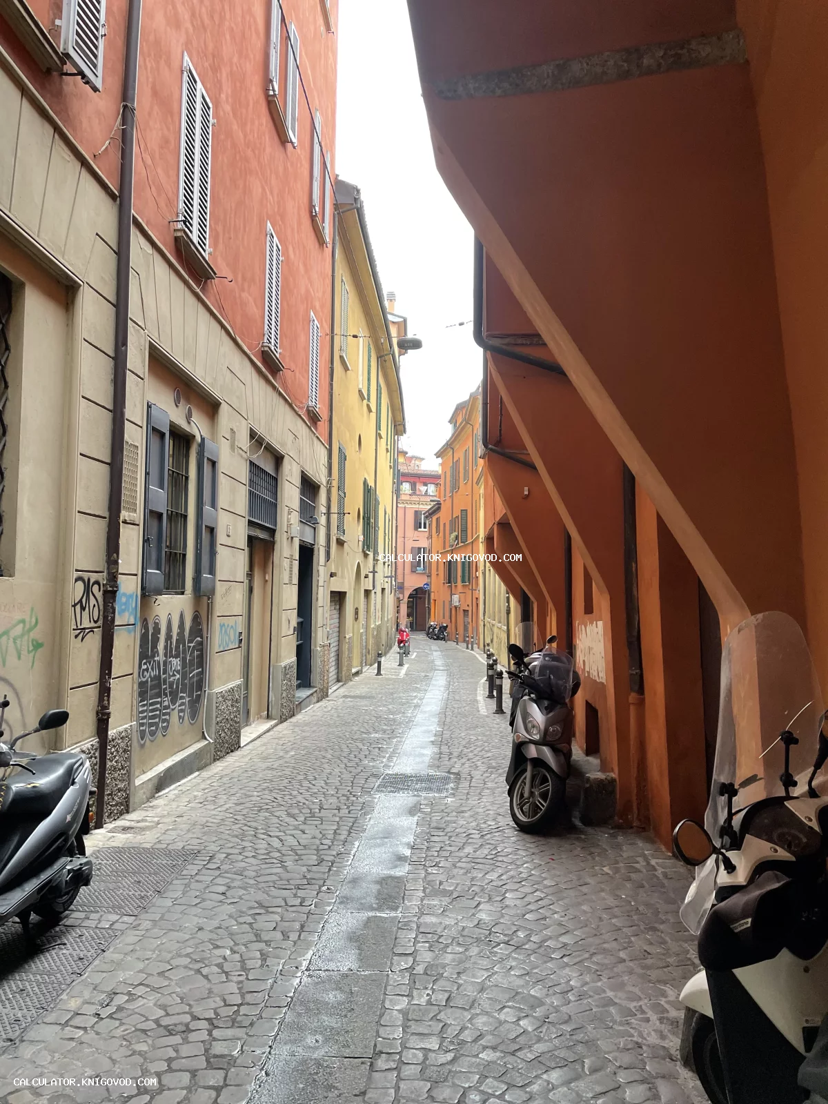 Узкая мощеная улица в европейском городе с высокими зданиями теплых оттенков и припаркованными скутерами.