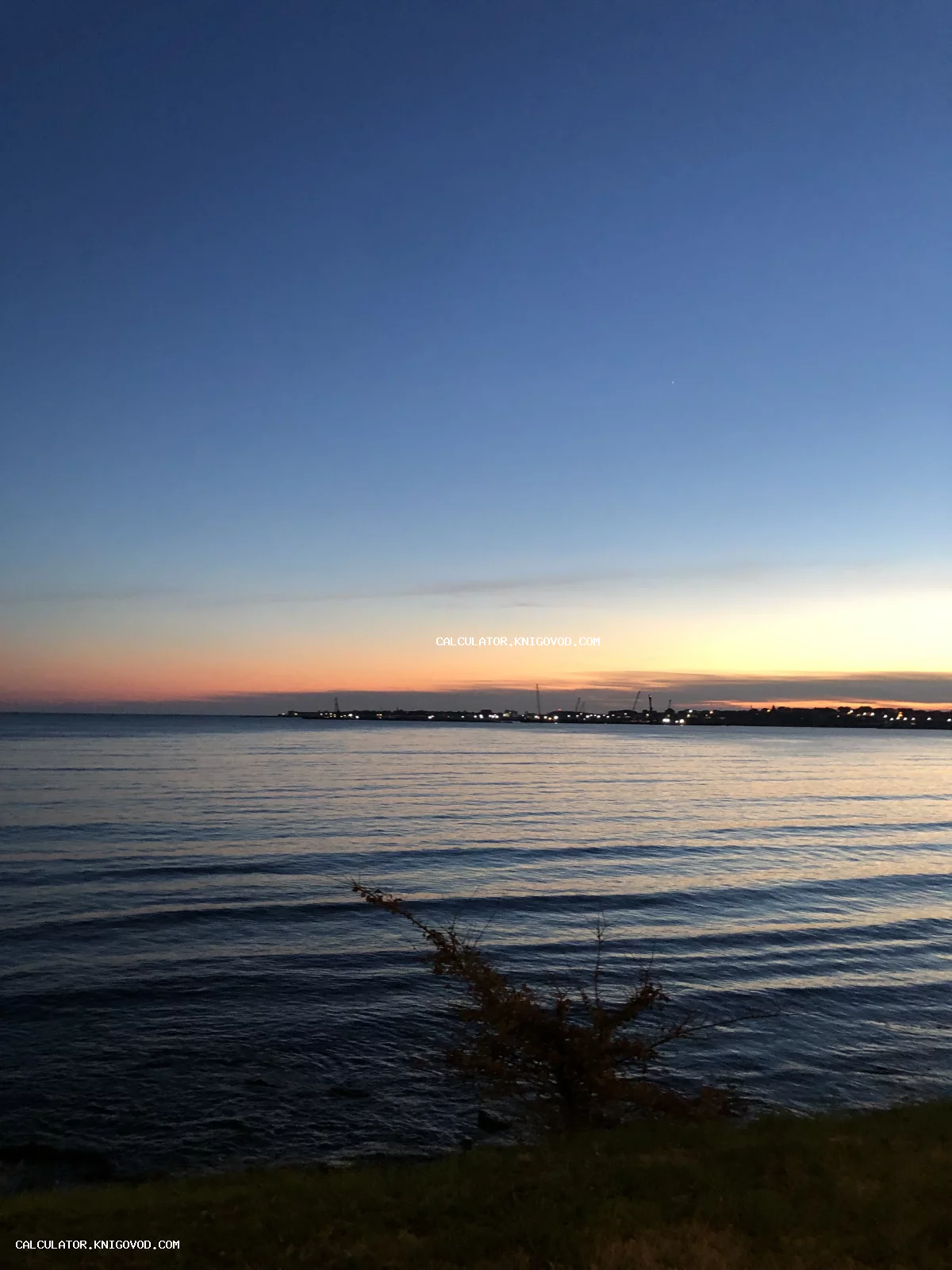 Пейзаж с вечерним закатом над морем, огни порта на горизонте и темная вода с рябью.