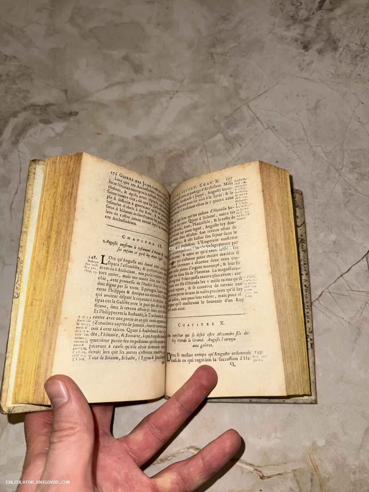 Раскрытая старинная книга с пожелтевшими страницами и французским текстом, удерживаемая рукой на фоне мраморной поверхности.