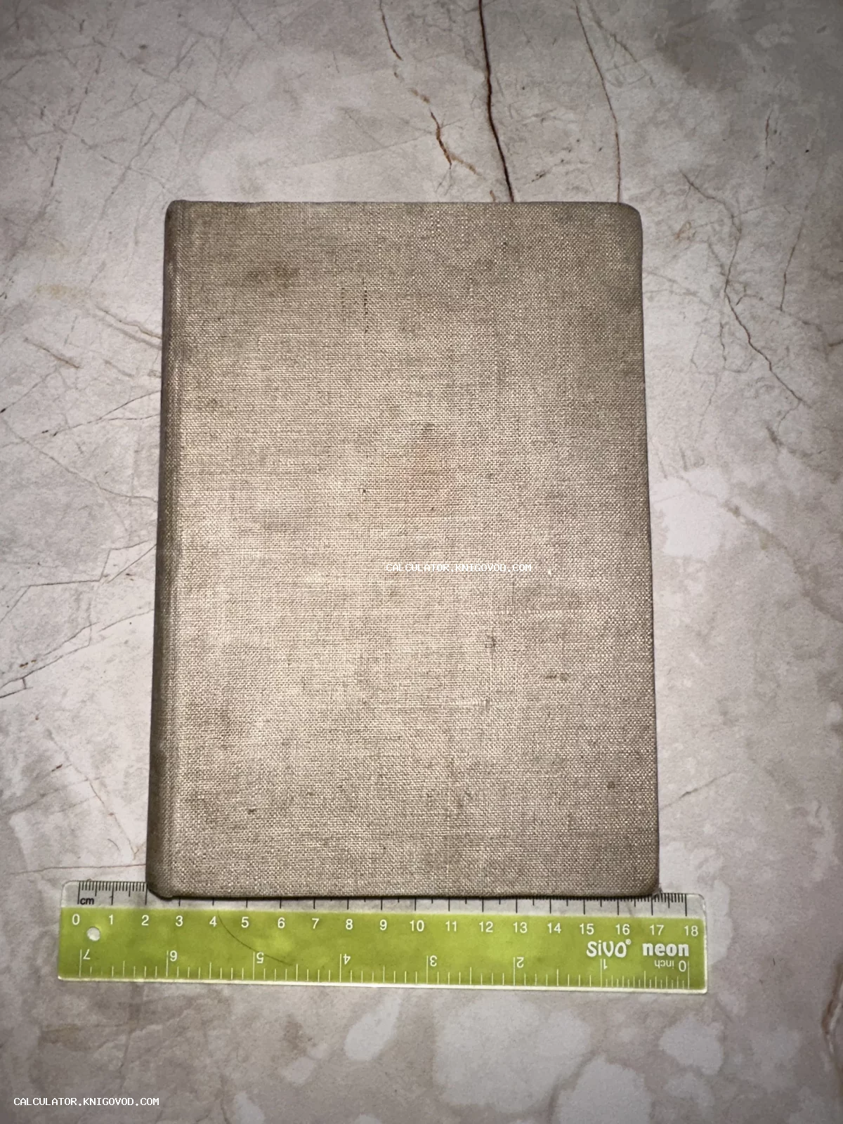 Старинная книга в сером льняном переплете лежит на мраморной поверхности рядом с линейкой.