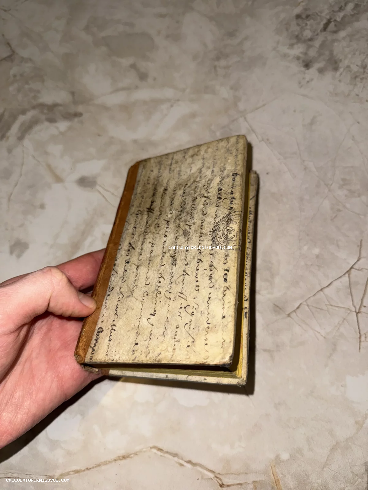 Маленькая старинная книга в кожаном переплете, обложка которой украшена рукописным каллиграфическим текстом и печатью.