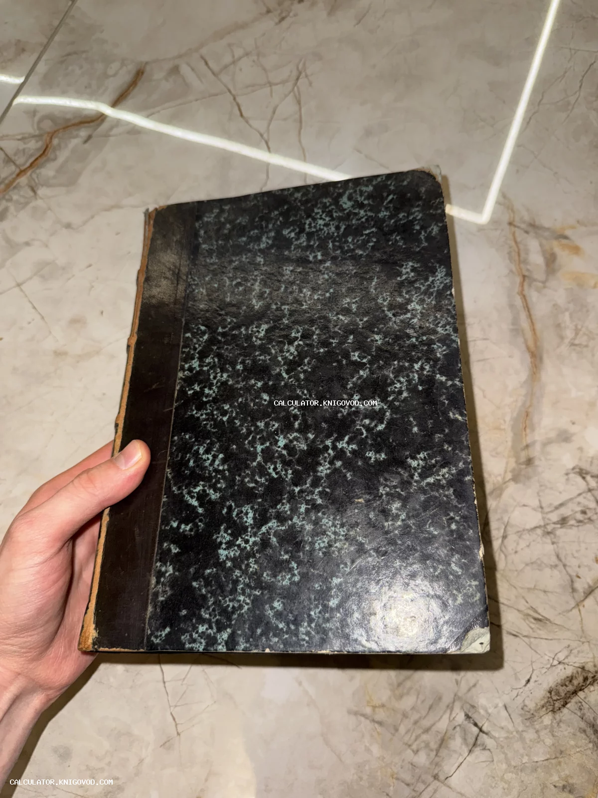 Старинная книга с темно-зеленым мраморным узором на обложке и потертым темным корешком в руке.