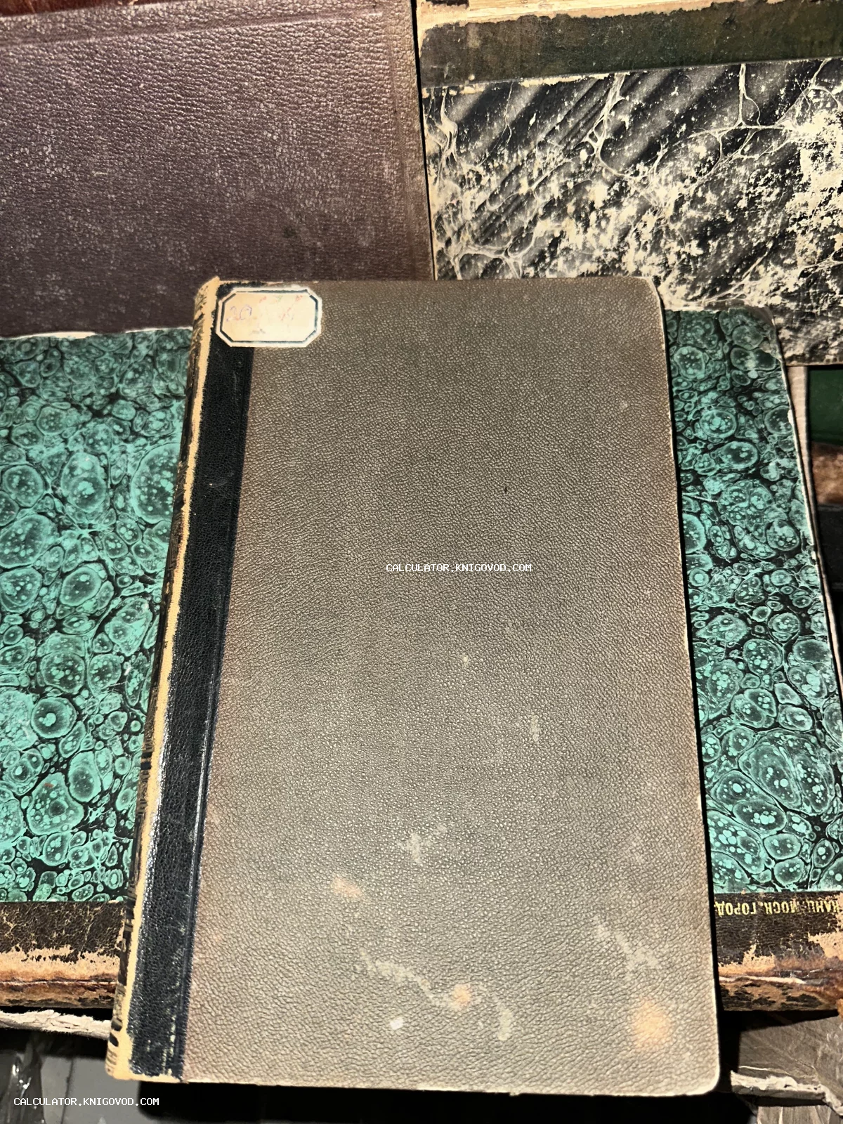 Антикварная книга с серой текстурированной обложкой и черным корешком на фоне других старых книг с мраморным узором.