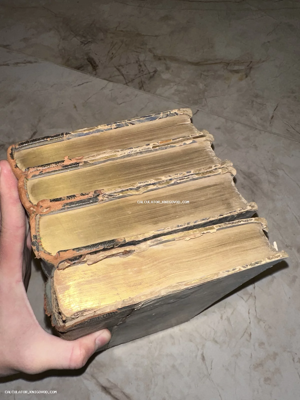 Четыре массивные старинные книги с позолоченными торцами страниц в руке человека на фоне мраморного пола.