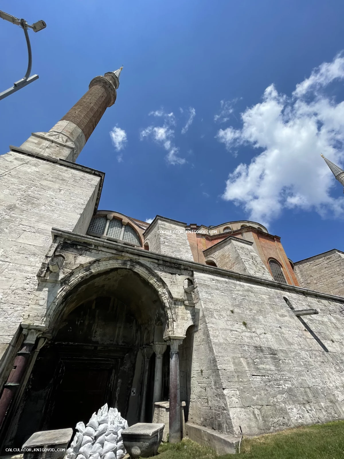 Вид снизу на массивные каменные стены, входную арку и минарет собора Святой Софии в Стамбуле на фоне ярко-синего неба с облаками.