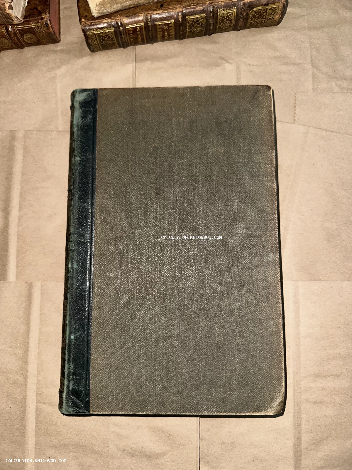 Старинная книга в сером тканевом переплете с темным кожаным корешком, вид сверху на фоне упаковочной бумаги.