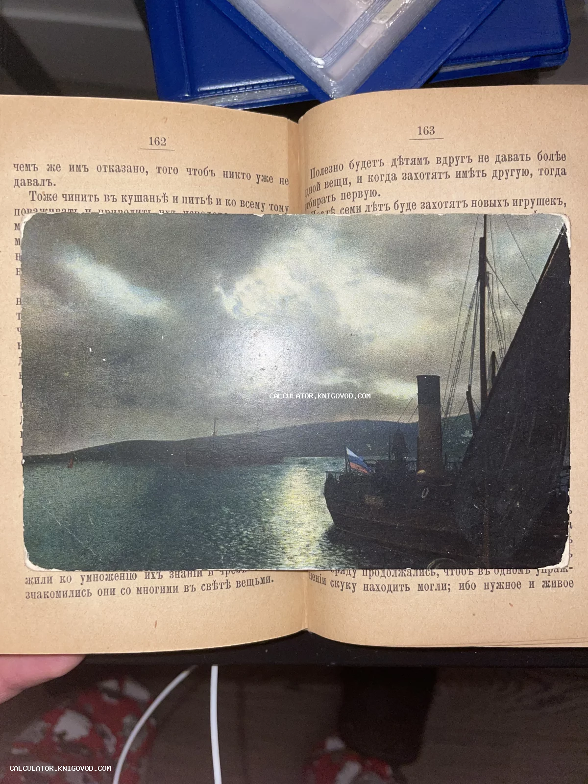 Старинная цветная открытка с изображением корабля на фоне закатного моря, лежащая на страницах раскрытой дореволюционной книги.