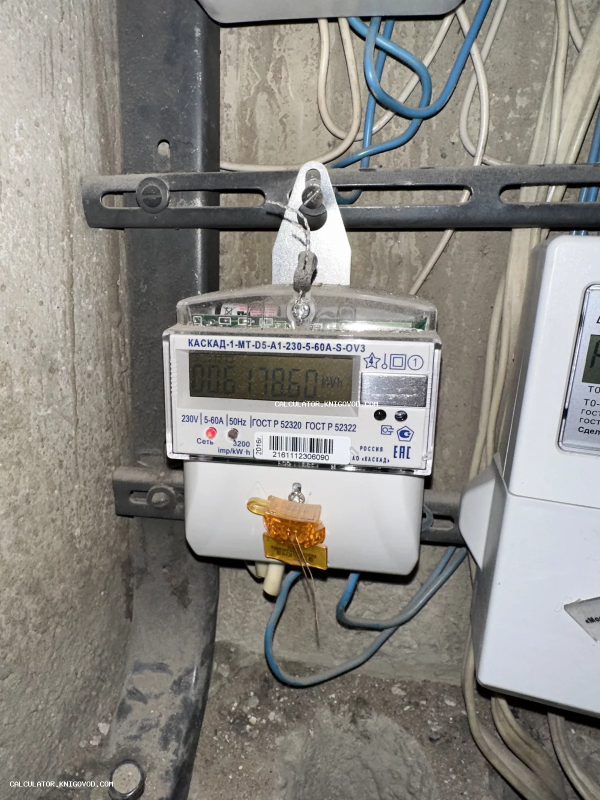 Современный многотарифный электросчетчик Каскад с цифровым табло и защитной пломбой, установленный на бетонной стене среди проводов.