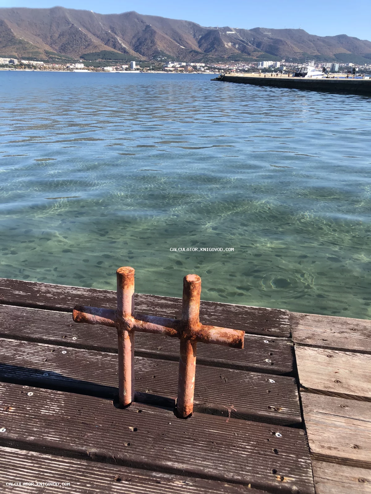 Старый ржавый кнехт на деревянном настиле пирса на фоне прозрачного моря и гор.