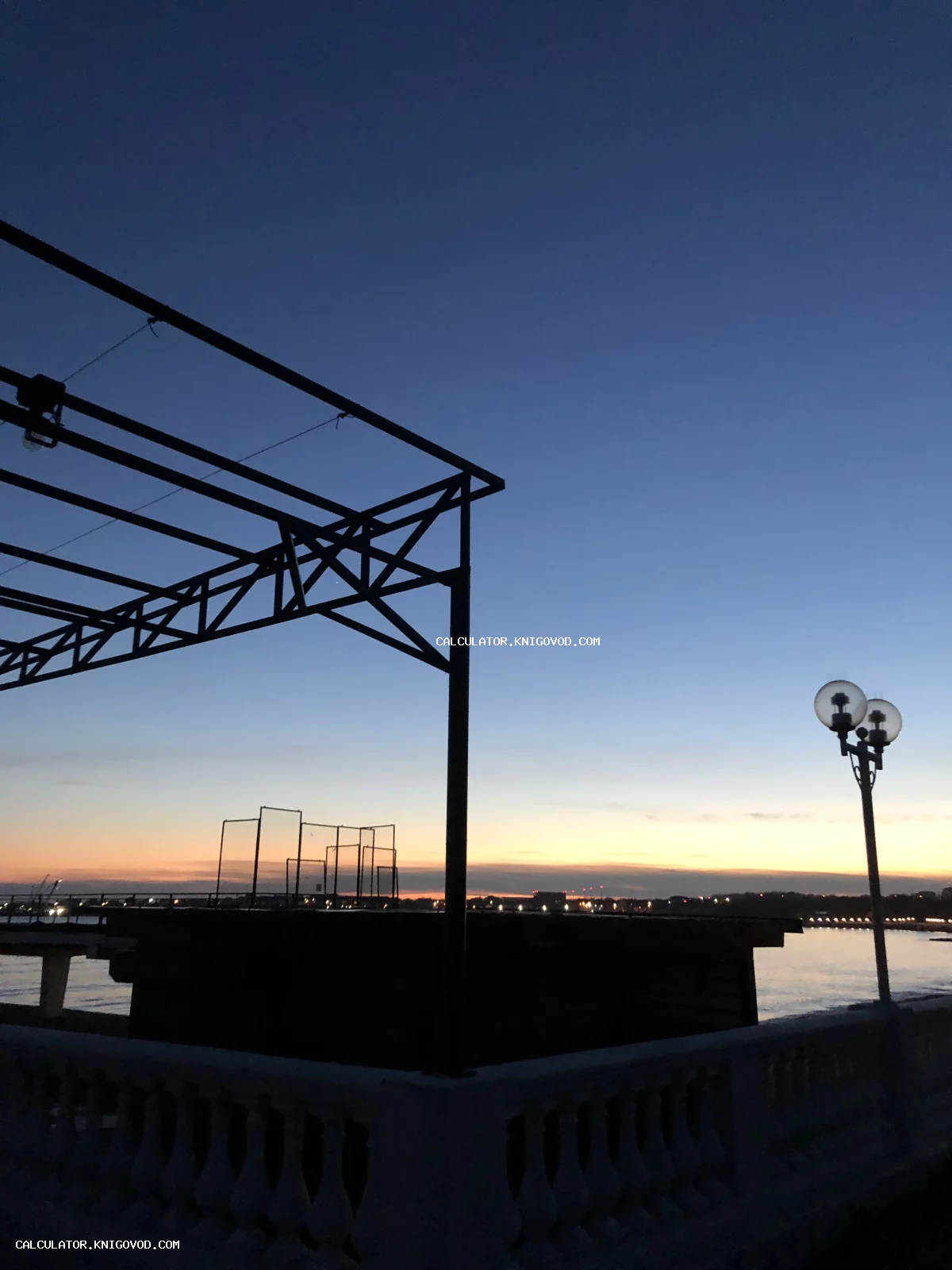 Силуэты металлических конструкций и уличного фонаря на фоне оранжевого заката над рекой.