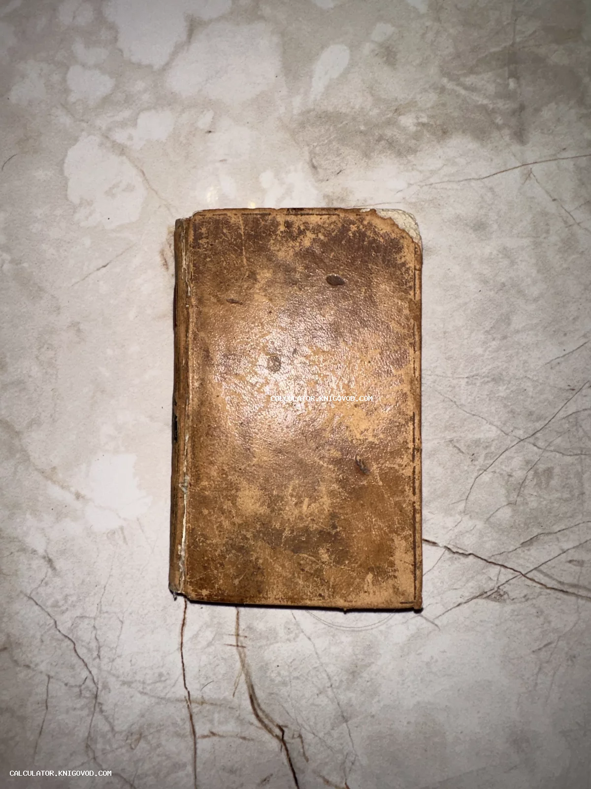 Вид сверху на старинную книгу малого формата в потертом коричневом кожаном переплете на светлом мраморном фоне.